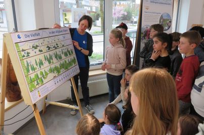 Eine Gruppe von Kindern hört aufmerksam zu, während ein Lehrer an einem Diagramm erklärt. Im Hintergrund sind Fenster und weitere Informationsmaterialien zu sehen.