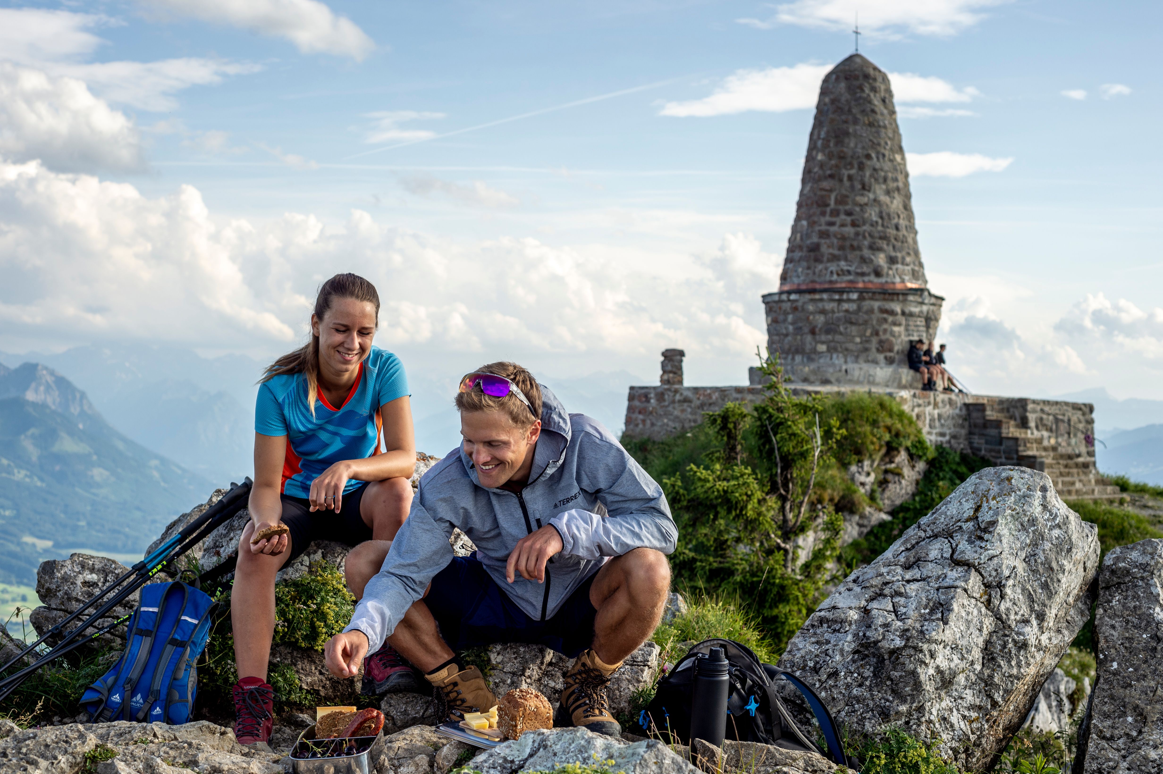 Zwei Wanderer genießen die Brotzeit auf dem Gipfel des 1.738 m hohen Grüntens. Im Hintergrund sind ist das Jägerdenkmal und die Allgäuer Hochalpen zu sehen.