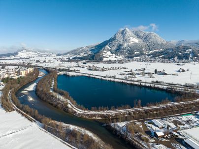 Eine schneebedeckte Landschaft mit einem klaren blauen See und Bergen im Hintergrund. Der Fluss schlängelt sich durch die weiße Winterlandschaft.