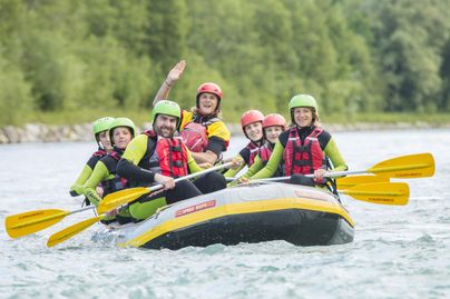 Eine Gruppe von Menschen paddelt fröhlich in einem Raft auf einem Fluss. Sie tragen Schutzhelme und Schwimmwesten und genießen ein aufregendes Abenteuer in der Natur.