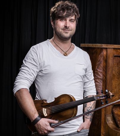 Ein Mann steht mit einer Violine in der Hand vor einem Klavier. Er trägt ein weißes Shirt und hat einen lässigen Ausdruck.