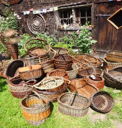 Eine Ansammlung von geflochtenen Körben in verschiedenen Größen liegt auf dem Gras. Im Hintergrund sind alte Holzwände und Blumen zu sehen.