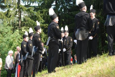 Eine Gruppe von Männern in historischen Uniformen mit hohen Hüten steht in einer gepflasterten Umgebung. Im Hintergrund sind Menschen und Bäume zu sehen.