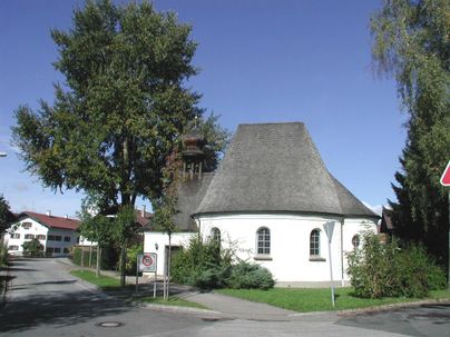 Eine kleine Kirche mit einem runden Dach und einer schönen Umgebung. An der Straße stehen Bäume und Wohngebäude.