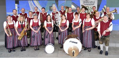 Eine Musikergruppe in traditionellen Trachten steht zusammen. Sie halten verschiedene Instrumente und posieren vor einer farbigen Wand.