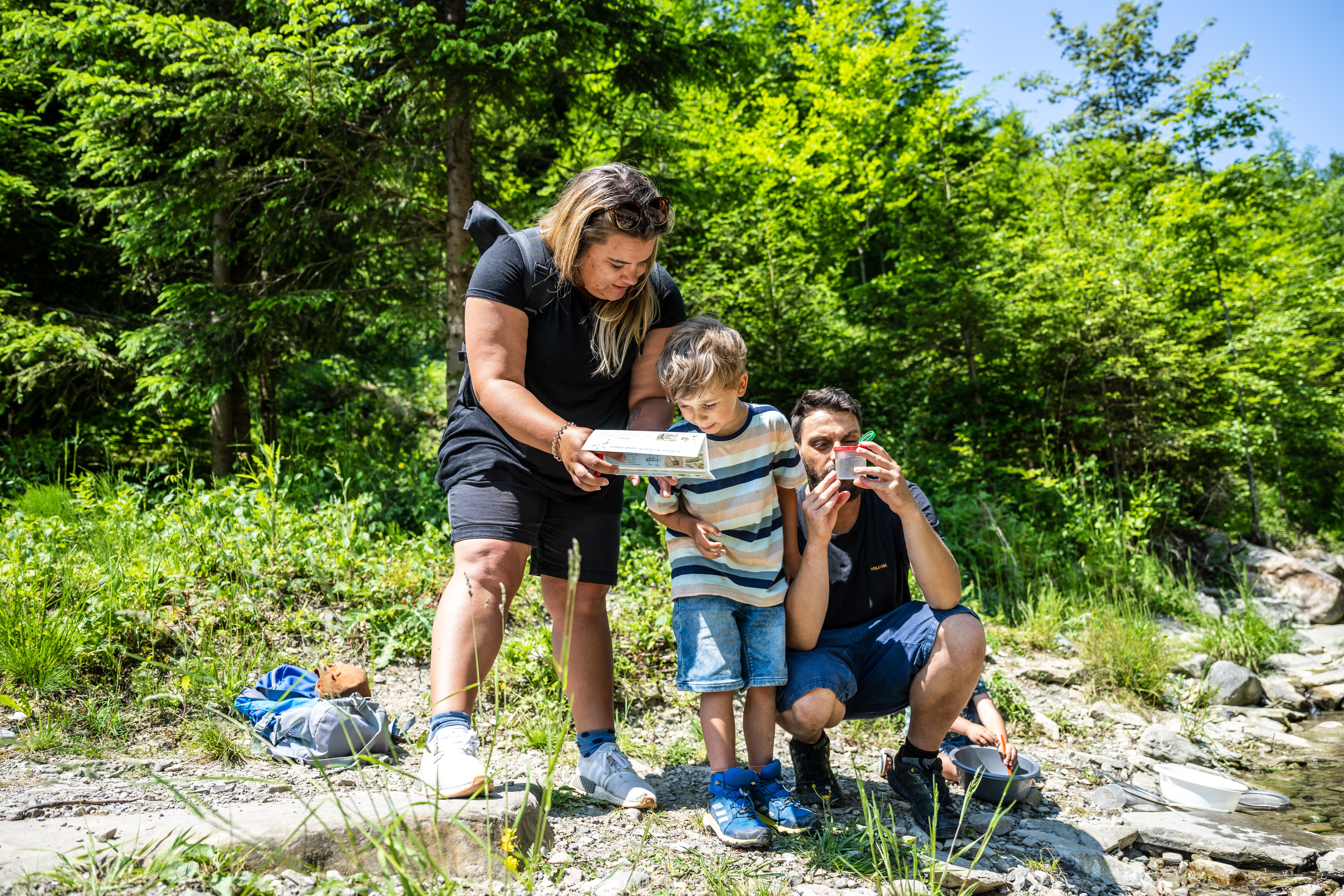 Eine Familie testet am Galetschbach den Forscherrucksack für den Erlebnispfad. Während sich der Vater mit der Becherlupe beschäftigt, studieren Mutter und Sohn den beigelegten Kosmo-Natur-Führer.