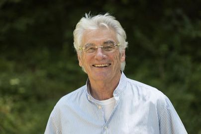 Ein älterer Mann mit grauen Haaren und einer Brille lächelt freundlich. Er trägt ein gestreiftes Hemd und steht vor einem grünen Hintergrund.
