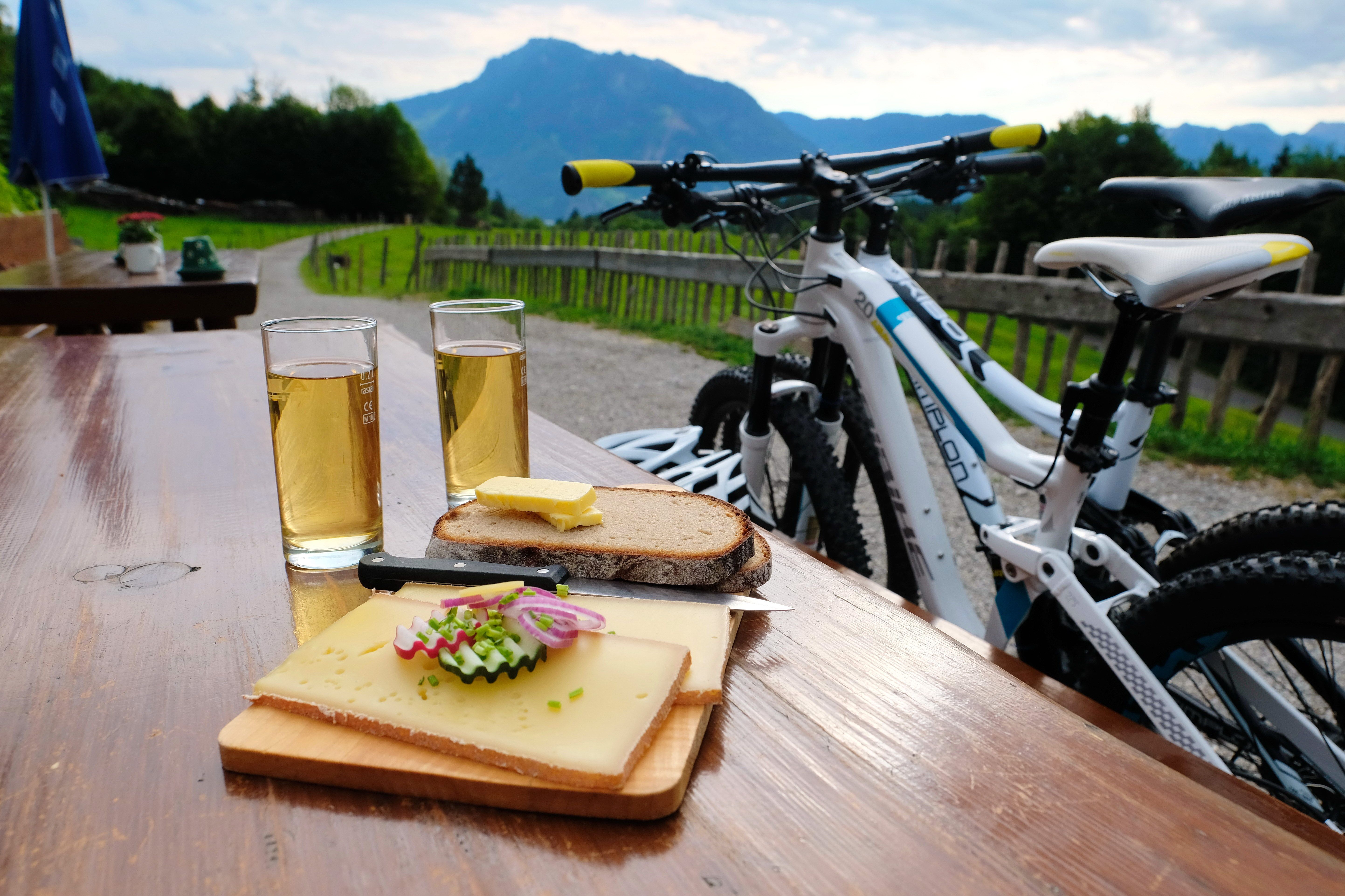 Zwei Getränke stehen gemeinsam mit einem Brotzeitbrett, mit Käse und Brot, auf einem Holztisch. Angelehnt an den Tisch stehen zwei Mountainbikes. Im Hintergrund sieht man den Grünten.