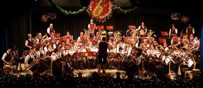 Ein Blasorchester spielt auf einer Bühne. Die Musiker tragen traditionelle Kleidung und sind konzentriert bei der Aufführung.
