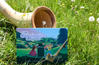 Ein Buch mit dem Titel "Das Geheimnis des Alphorns" liegt auf grünem Gras. Im Hintergrund ist ein Alphorn und eine idyllische Landschaft mit Kindern zu sehen.