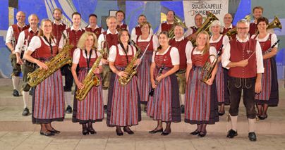 Eine Musikgruppe in traditioneller Tracht steht zusammen und hält Blasinstrumente. Sie sind in einem festlichen Ambiente und lächeln in die Kamera.