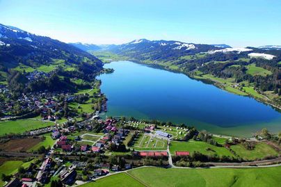Ein malerischer See umgeben von grünen Wiesen und Bergen. Die Landschaft ist sonnig und ruhig, ideal für einen Ausblick.