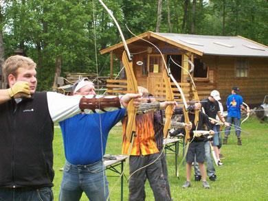 Eine Gruppe von Personen, die im Freien mit Bögen und Pfeilen schießt. Im Hintergrund sieht man eine Holzlaube und Bäume.