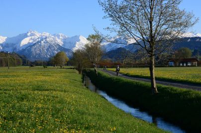 Wanderer und Radfahrer genießen eine Tour durch die Illerauen mit Blick in die verschneiten Oberstdorfer Berge.