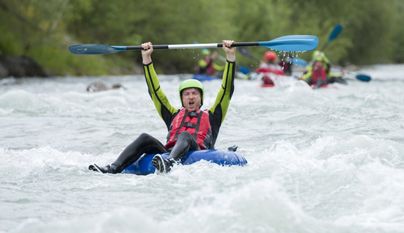 Ein Kajakfahrer hebt jubelnd das Paddel über den Kopf. Im Hintergrund sind weitere Paddler auf einem reißenden Fluss zu sehen.