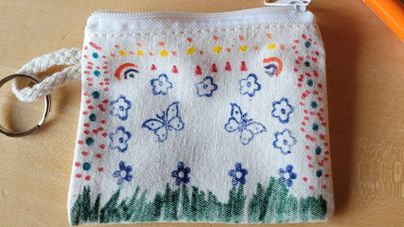 Eine Hand hält eine lila Perle an einem Faden. Daneben sind verschiedene Perlen und eine zusammengebaute Perlenkette zu sehen.