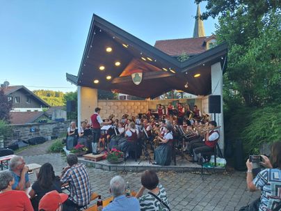 Ein Musikensemble spielt auf einer Freiluftbühne. Die Zuschauer sitzen entspannt und genießen die Darbietung in einer schönen Umgebung.