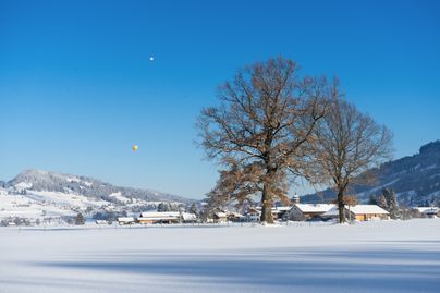 Der Burgberger Ortsteil Agathazell im Winter. Am Himmel schweben zwei Heißluftballons. Winterwanderer, Schneeschuhgeher und Langläufer kommen auf gut präparierten Wegen und Loipen in der Gemeinde Blaichach voll auf Ihre Kosten.