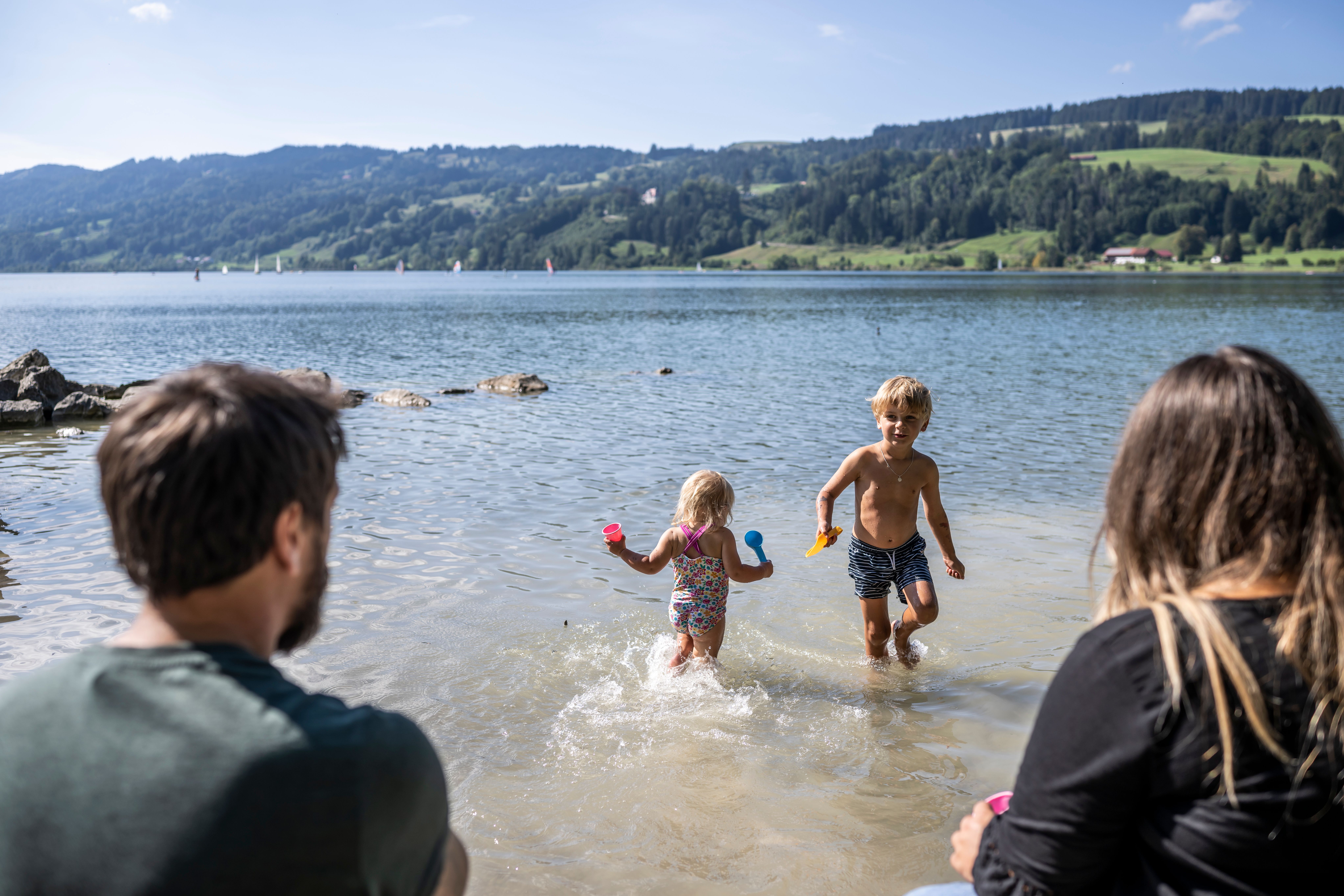 Zwei Kinder plantschen gemeinsam am Rand des Alpsees in Immenstadt. Ihre Eltern sitzen am Ufer.
