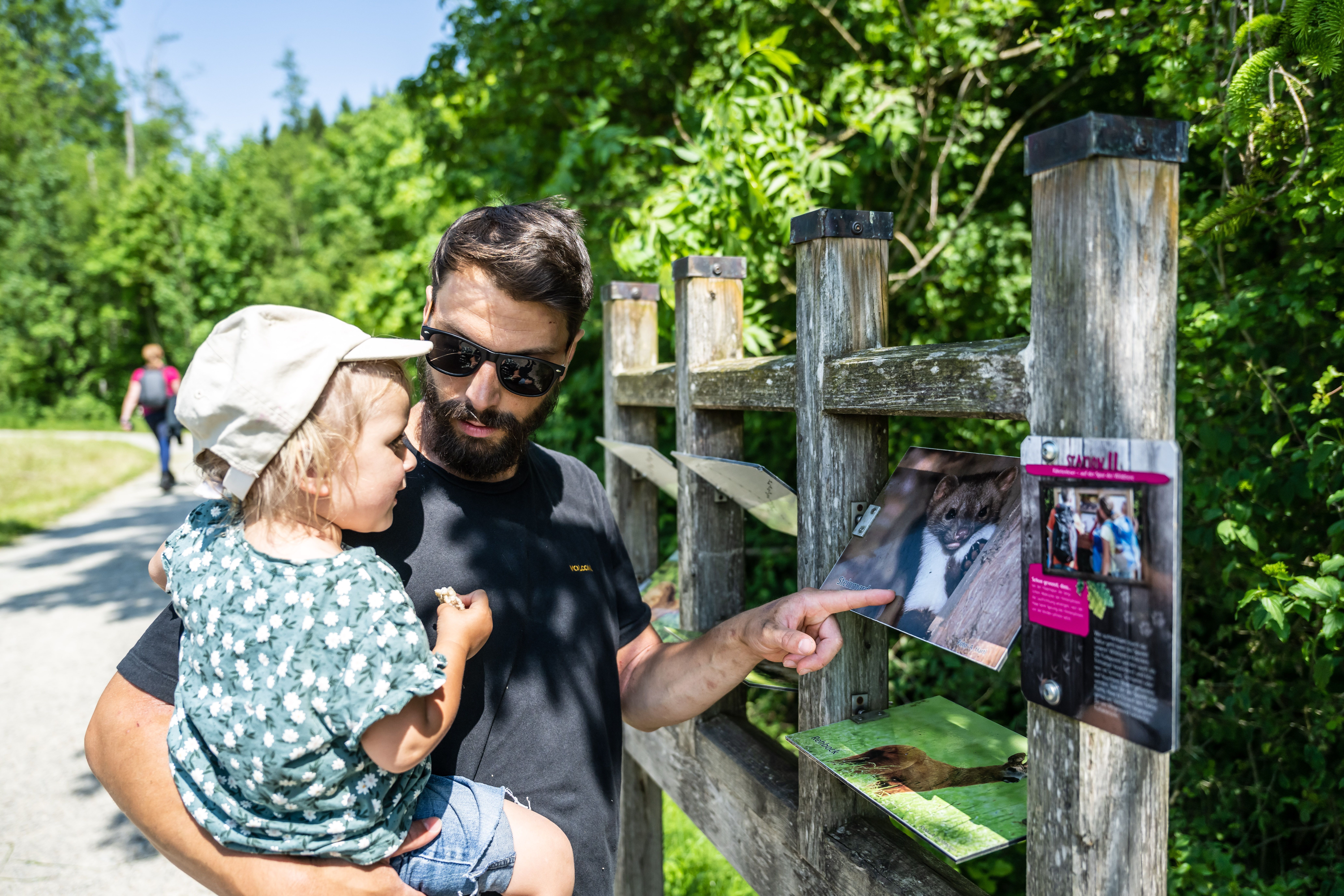 Ein Vater erklärt seiner Tochter auf dem Arm die Spuren der Tiere im Wald an der elften Erlebnisstation entlang des Elebnispfads Galetschbach, dem Memorie "Fährtenlesen - auf den Spuren der Wildtiere".