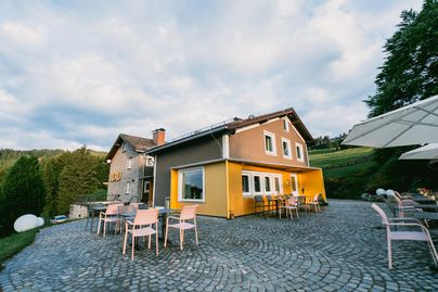 Ein modernes Gebäude mit einer gelben Fassade und einer Terrasse. Rund um das Haus stehen pinke Stühle und Tische auf einem gepflasterten Platz.