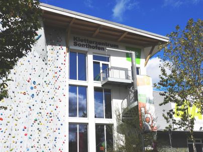 Ein modernes Kletterzentrum mit bunten Klettersockeln und großen Fenstern. Die Fassade ist hell und einladend, umgeben von Bäumen und einem blauen Himmel.