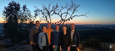 Eine Gruppe von sechs Personen steht zusammen in der Natur. Im Hintergrund ist ein schöner Sonnenuntergang und ein Baum mit Wimpeln zu sehen.