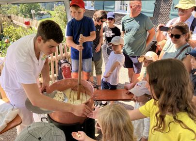 Ein junger Mann rührt in einem großen Topf, während eine Gruppe von Kindern und Erwachsenen zuschaut. Die Szene findet im Freien statt und vermittelt eine fröhliche Lernatmosphäre.