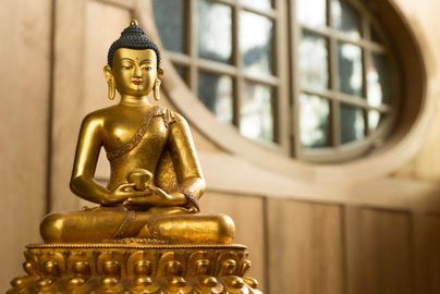 Eine goldene Buddha-Statue sitzt auf einem dekorativen Sockel. Im Hintergrund sind Holzdetails und runde Fenster sichtbar.