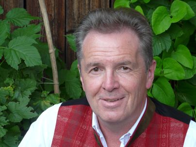 Ein Mann in traditioneller Tracht lächelt in die Kamera. Im Hintergrund sind grüne Pflanzen zu sehen.
