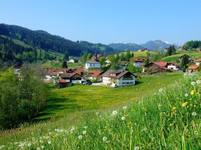 Eine malerische Landschaft mit einem kleinen Dorf und grünen Wiesen. Im Hintergrund sind Berge und ein blauer Himmel zu sehen.