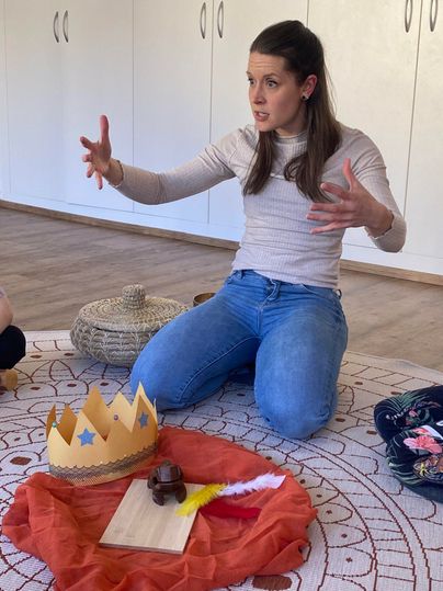 Eine Frau sitzt auf dem Boden und erzählt begeistert. Vor ihr liegen ein rotes Tuch, eine Krone und einige Gegenstände.