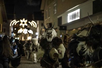 Eine Gruppe von Menschen in traditionellen Kostümen zieht durch die Straßen bei Nacht. Lichter funkeln über ihnen und schaffen eine festliche Atmosphäre.