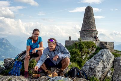 Zwei Wanderer genießen die Brotzeit auf dem Gipfel des 1.738 m hohen Grüntens. Im Hintergrund sind ist das Jägerdenkmal und die Allgäuer Hochalpen zu sehen.