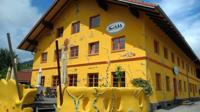 Ein auffälliges, gelbes Gebäude mit bunten Wandmalereien. Vor dem Gebäude gibt es einen kleinen Außenbereich mit Terrassenmöbeln.