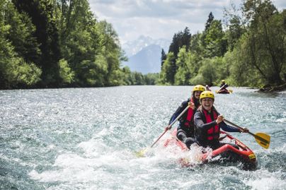 Eine Gruppe von Personen paddelt fröhlich in einem Schlauchboot auf einem klaren Fluss. Im Hintergrund sind grüne Bäume und Berge zu sehen.