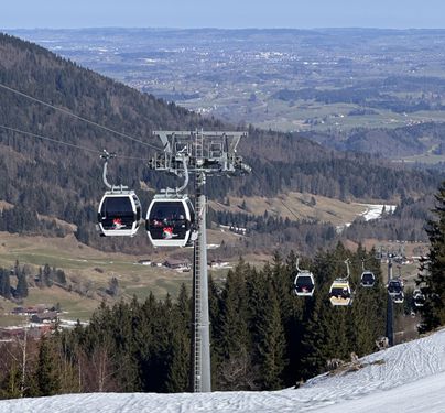 Eine Seilbahn fährt über schneebedeckte Berge. Im Hintergrund erstreckt sich das Tal mit Wäldern und Wiesen.