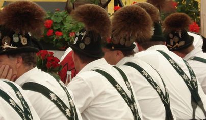 Eine Gruppe von Männern mit Trachtenhüten und traditionellen Lederhosen sitzt an einem Tisch. Im Hintergrund sind rote Blumen und Dekorationen sichtbar.