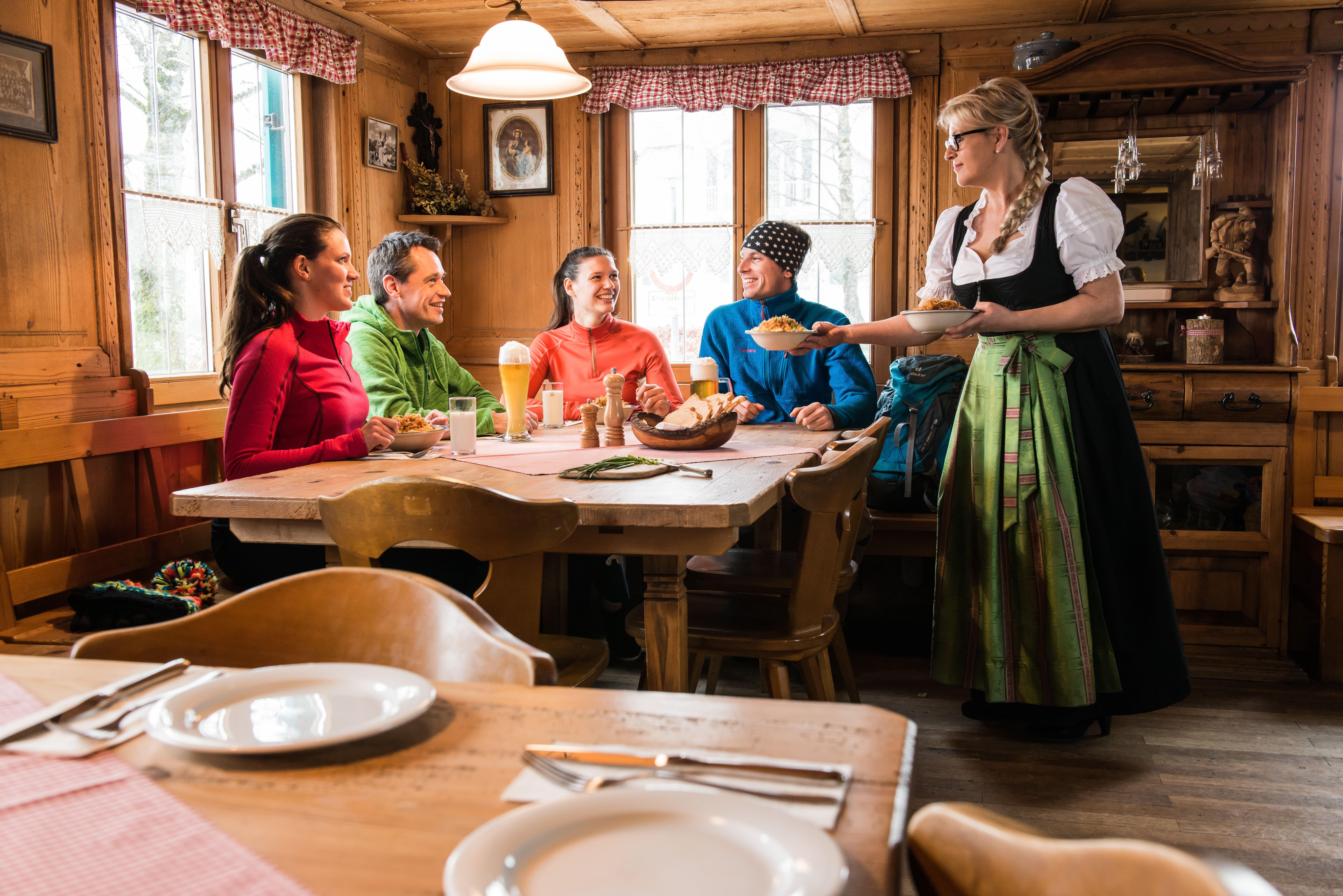 Eine Gruppe aus Leuten kehr im Gasthaus zum Lustigen Hirsch in Akams ein und wird von einer Frau in Tracht bedient.