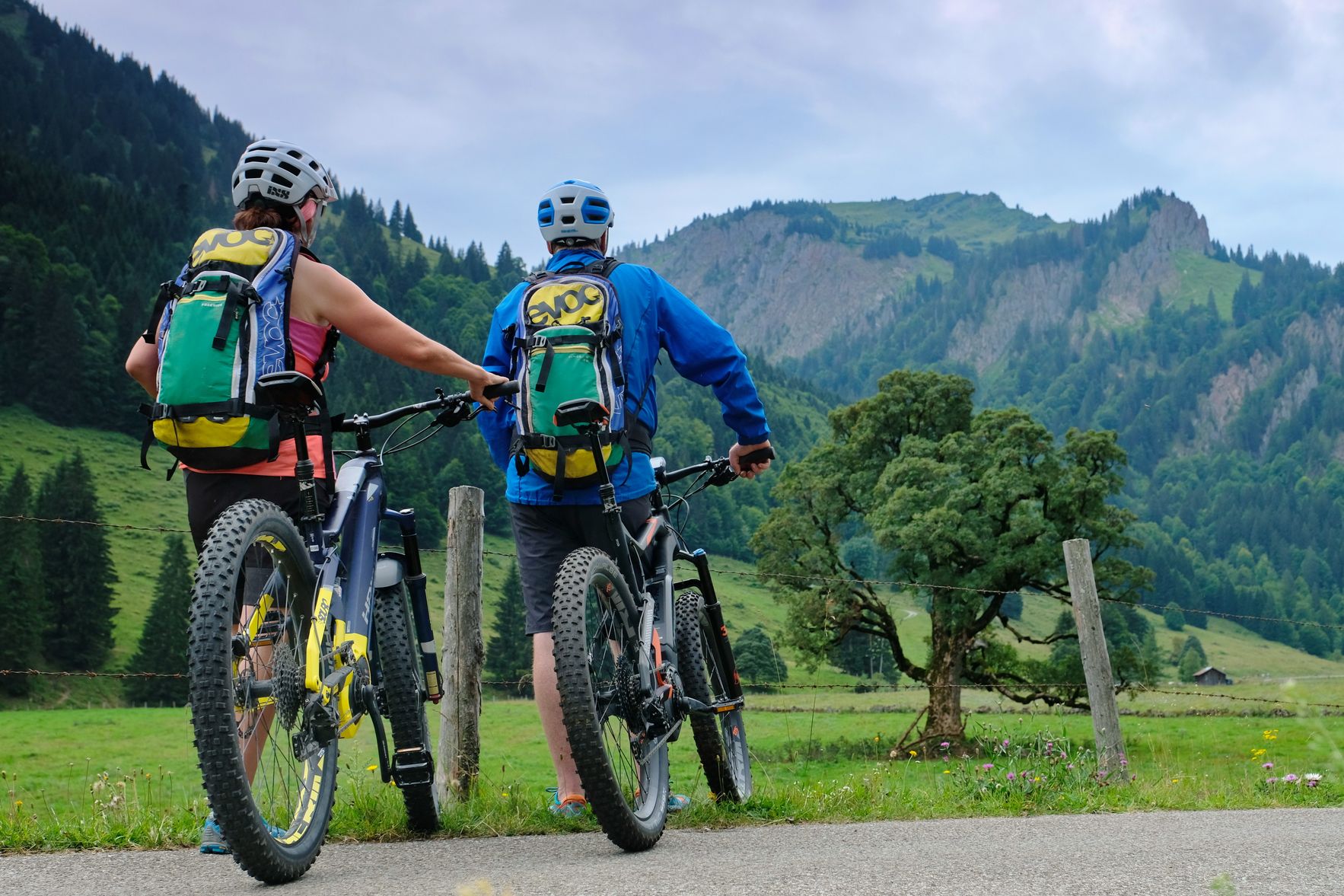 Zwei Mountainbiker stehen mit ihren E-Bikes am Stacheldrahtzaun einer Weide. Sie blicken in Richtung eines Berges im Gunzesrieder Tal.