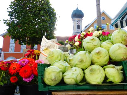 Ein bunter Marktstand mit frischem Gemüse, darunter viele Kohlköpfe. Im Hintergrund sind blühende Blumen und eine Kirche zu sehen.
