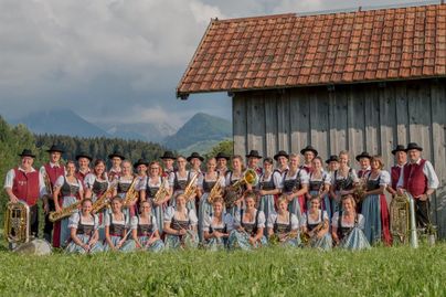 Eine Gruppe von Musikern in traditioneller Tracht steht vor einem Holzgebäude in einer ländlichen Umgebung. Sie halten verschiedene Musikinstrumente wie Trompeten und Tuben in den Händen.