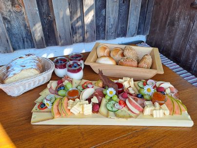 Ein appetitliches Brett mit Wurst, Käse und Essiggurken. Im Hintergrund ein Korb mit Brot und ein kühles Getränk.