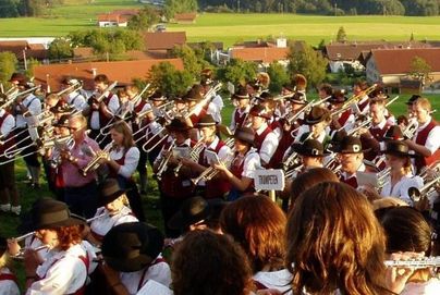 Eine Gruppe von Musikern spielt Trompeten und andere Blasinstrumente im Freien. Im Hintergrund sind grüne Wiesen und Fachwerkhäuser zu sehen.