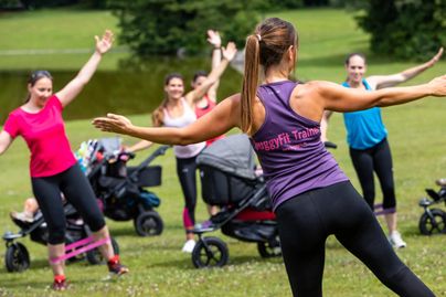 Eine Gruppe von Frauen trainiert im Freien mit Kinderwagen. Die Übungseinheit findet in einem grünen Park statt und zeigt ein aktives und geselliges Ambiente.
