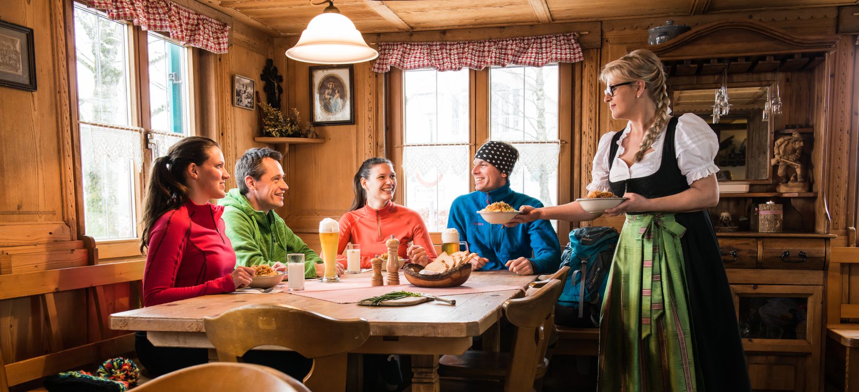 Eine Gruppe aus Leuten kehr im Gasthaus zum Lustigen Hirsch in Akams ein und wird von einer Frau in Tracht bedient.