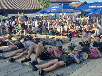 Eine fröhliche Menschenmenge beobachtet einen Wettkampf, bei dem Teilnehmer auf Holzstämmen liegen. Die Veranstaltung findet im Freien unter Sonnenschirmen statt.
