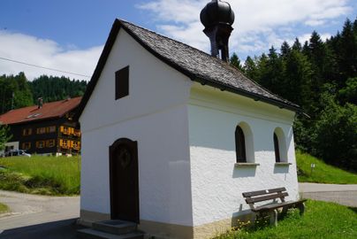 Eine kleine, weiße Kapelle mit einem dunklen Dach steht in einer grünen Umgebung. Im Hintergrund sind mehrere Holzhäuser zu sehen.