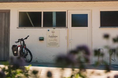 Ein Fahrrad steht an der Wand eines Gebäudes mit einem Schild, das Informationen enthält. Die Umgebung ist ruhig und freundlich gestaltet.