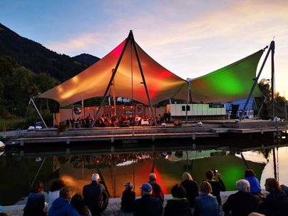 Ein Konzert unter einem großen Zelt mit bunten Lichtern spiegelt sich im Wasser. Viele Zuschauer sitzen am Ufer und genießen die Aufführung.